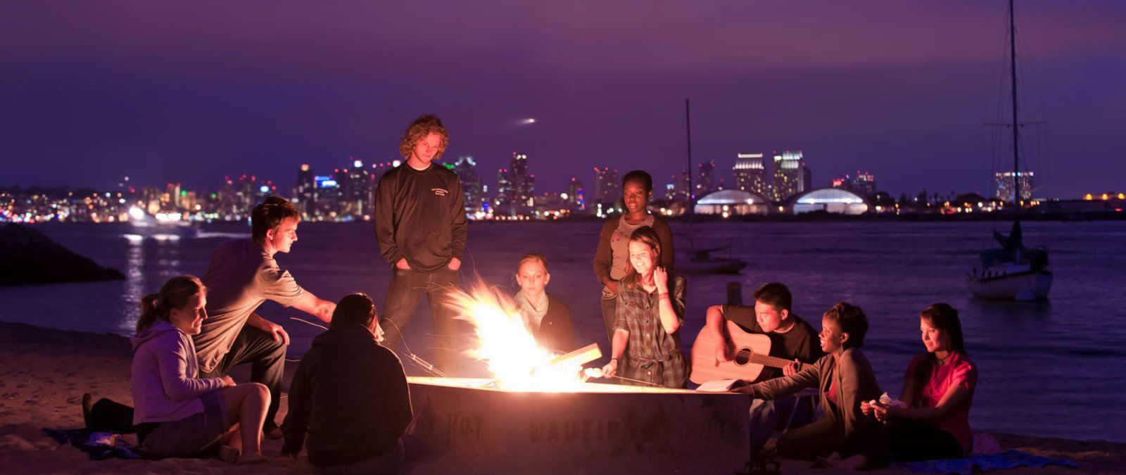 69 students have a beach bonfire at night and play music while at Shelter Island in San Diego.